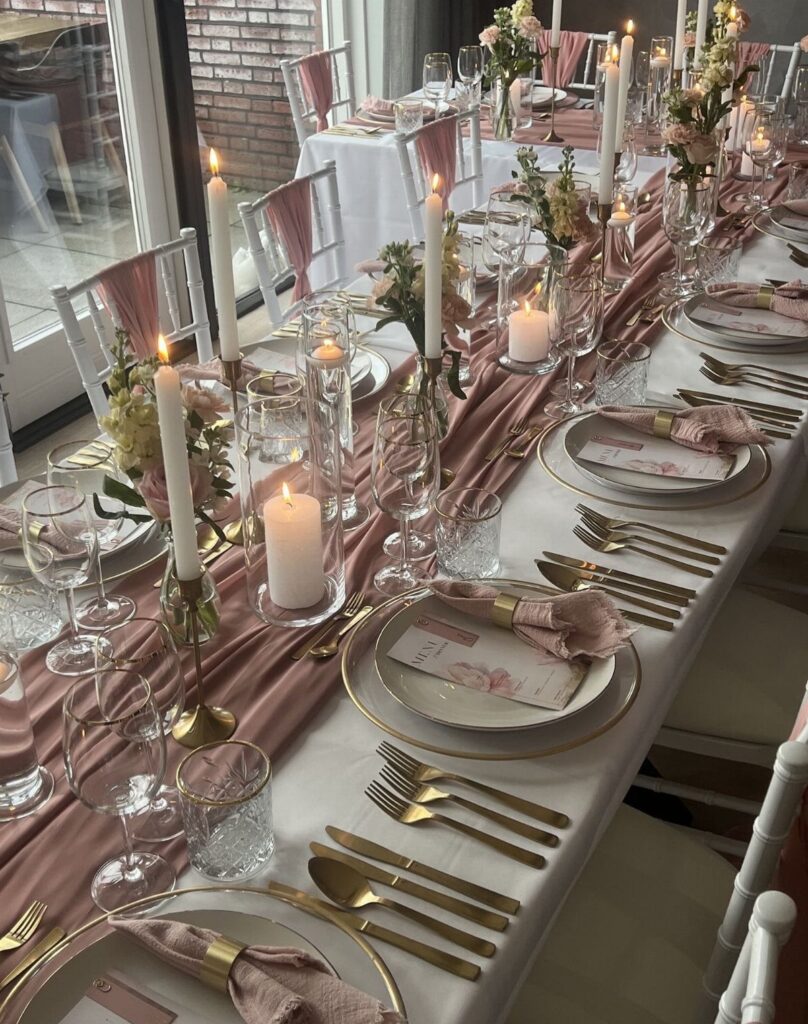 Elegant table setting with gold cutlery, rose runner, and tall white taper candles.