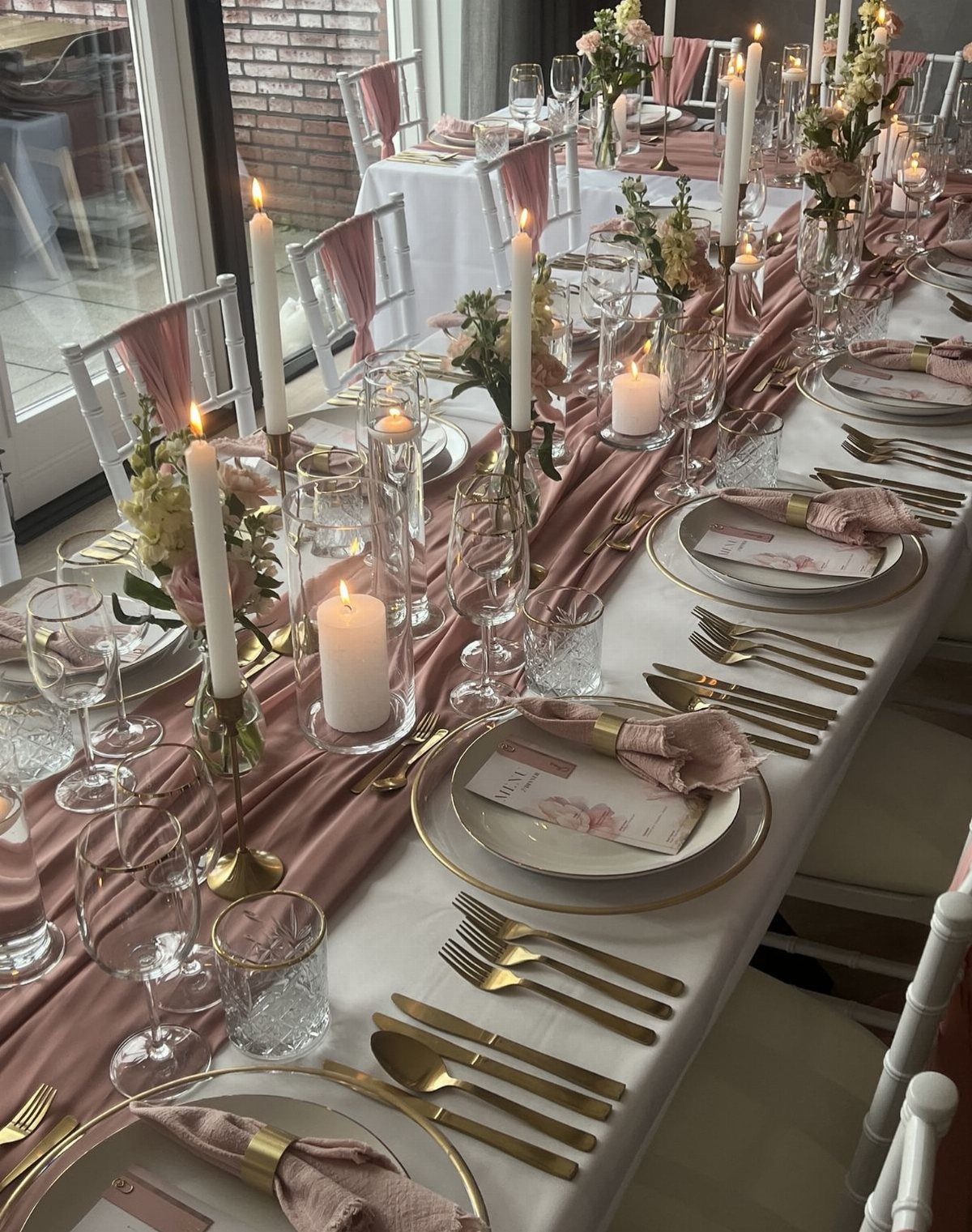 Elegant table setting with gold cutlery, rose runner, and tall white taper candles.