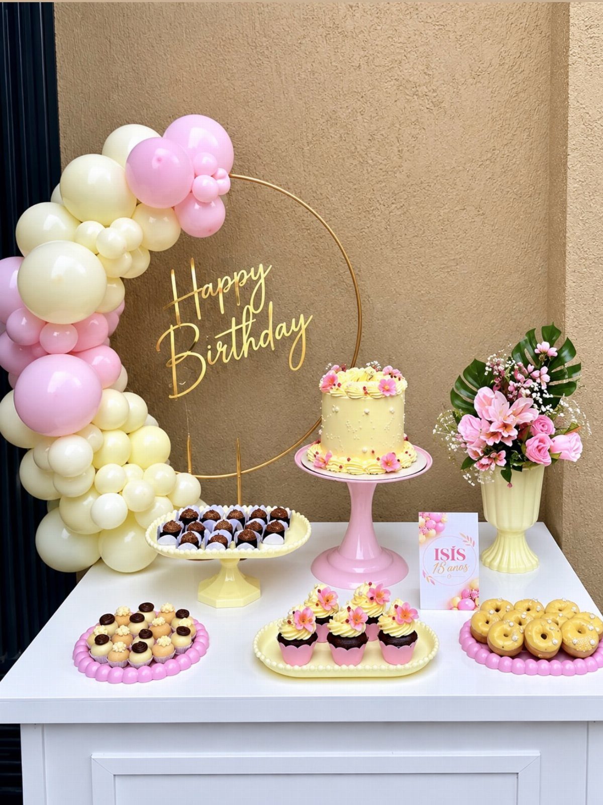 Birthday table setup with yellow cake, sweets, pink and yellow balloons, and flowers.