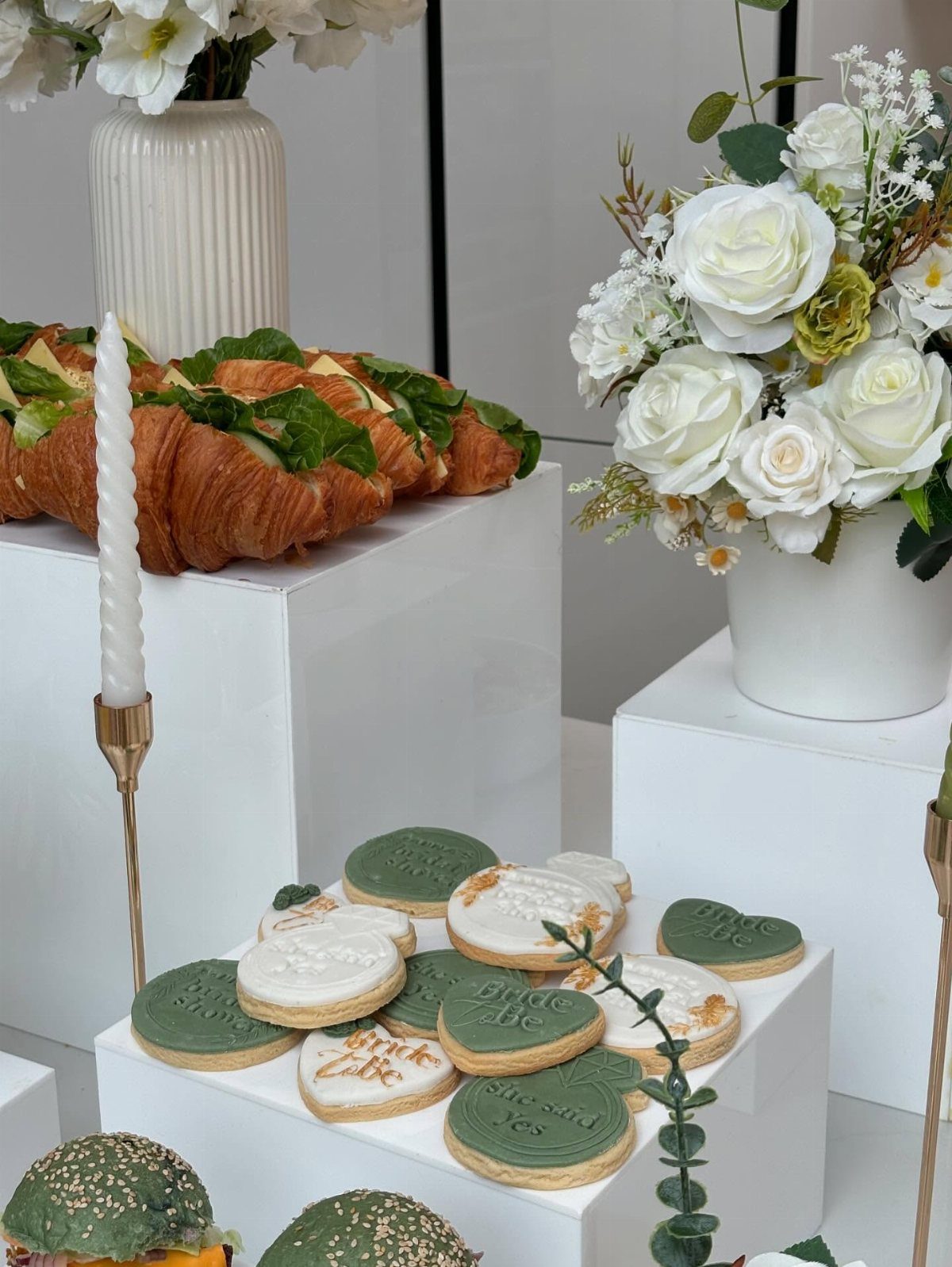 Elegant food display with frosted cookies, croissant sandwiches, and white roses on pedestals.