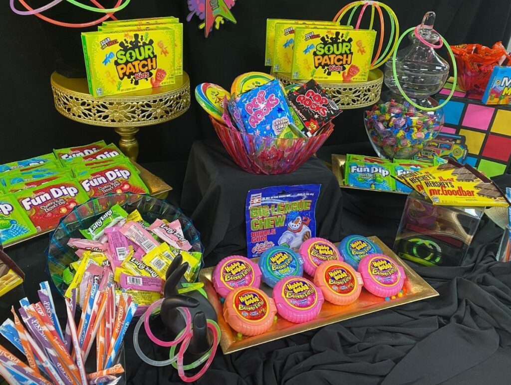 Colorful candy table display with various sweets, gold stands, and neon glow sticks.