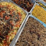 Aluminum foil trays filled with pasta, glazed meat, and rice for catering.