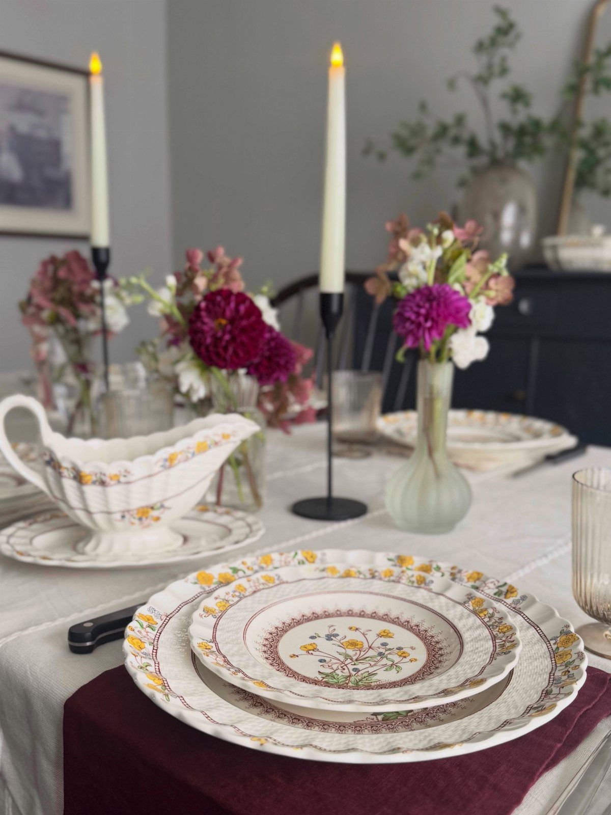 Floral patterned plates and purple flowers arranged on a dinner table with candles.