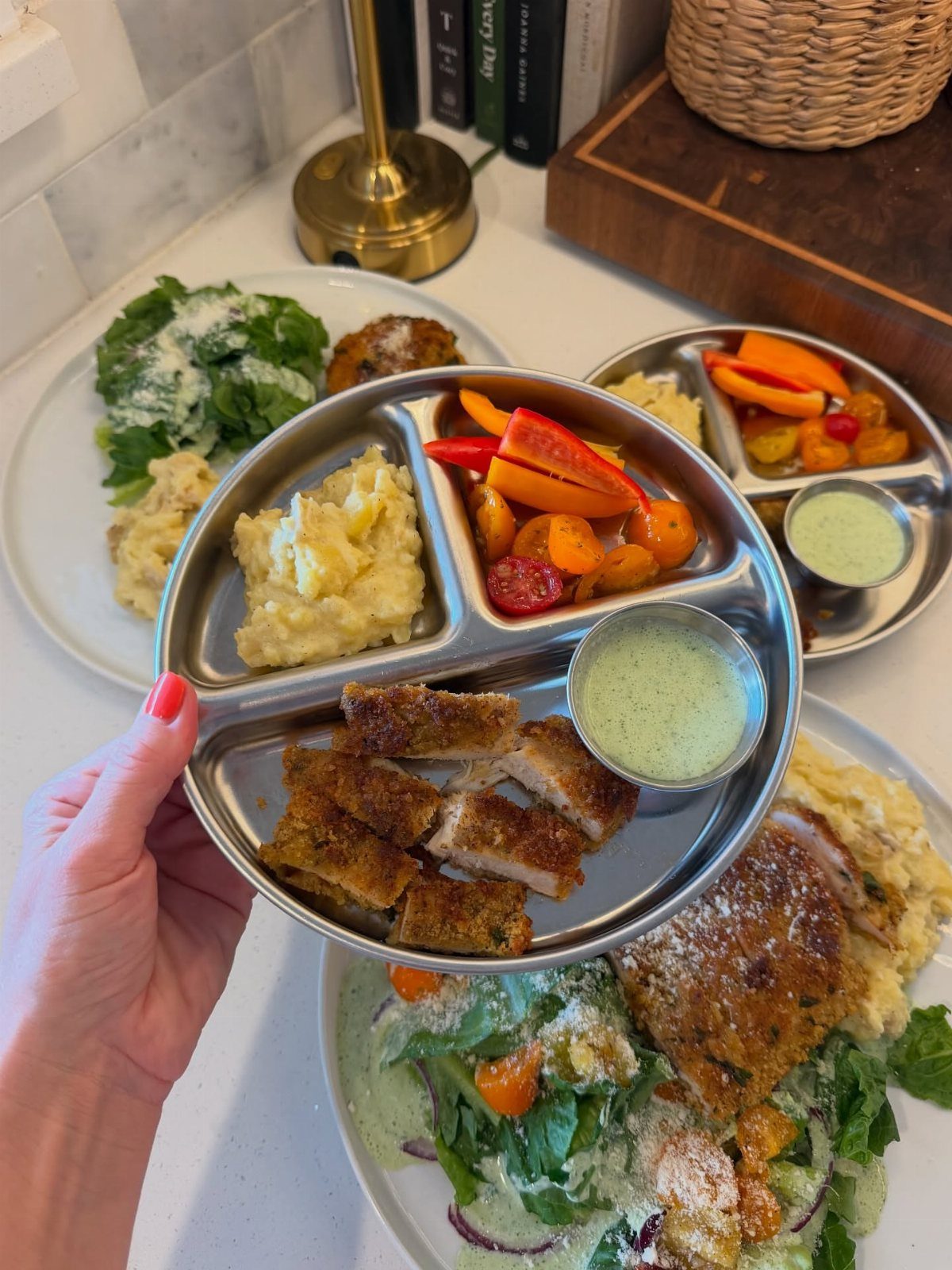 Divided metal tray with breaded chicken, mashed potatoes, and roasted peppers on countertop