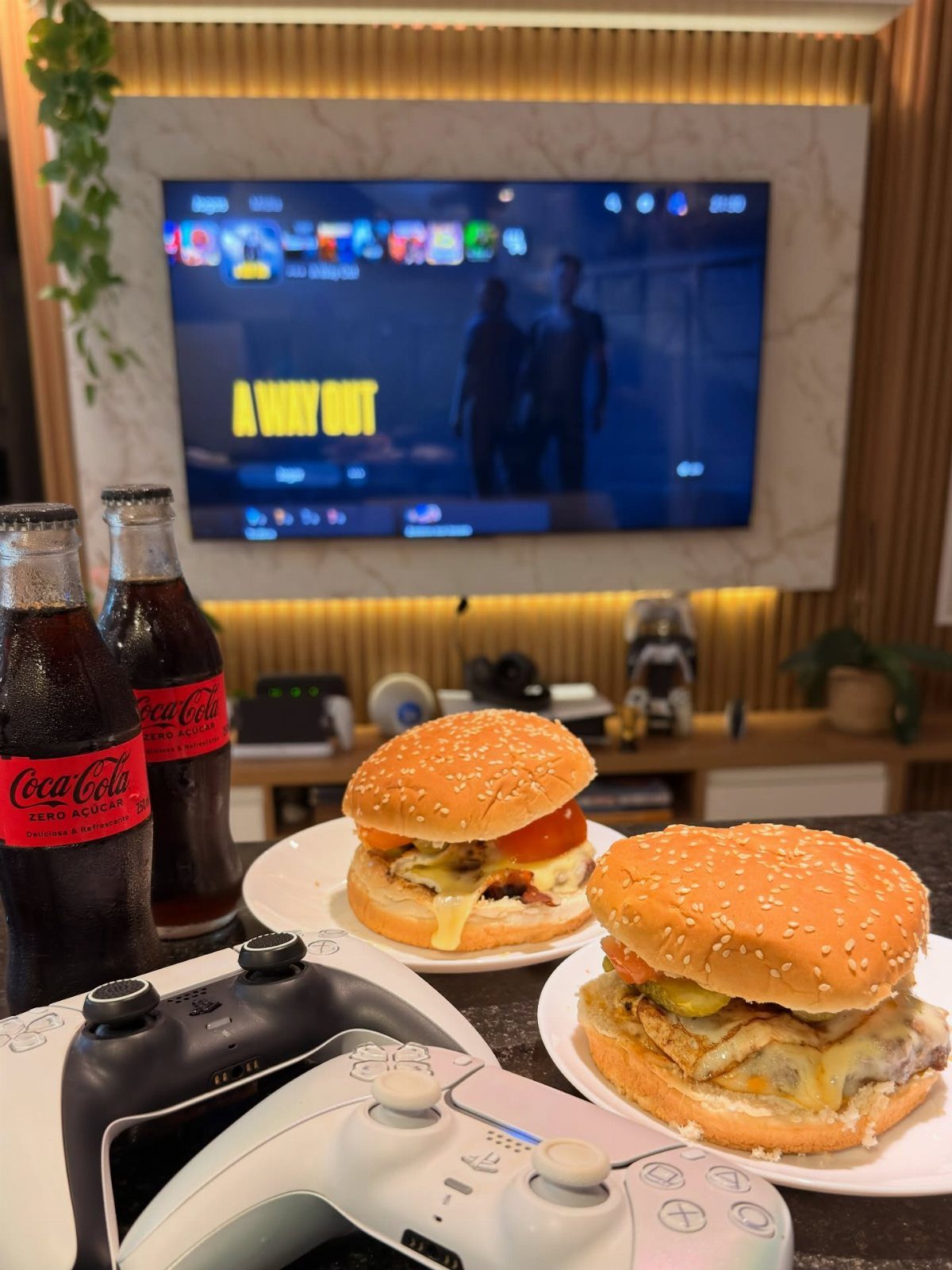Two cheeseburgers and Coca-Cola bottles next to game controllers before a television screen.