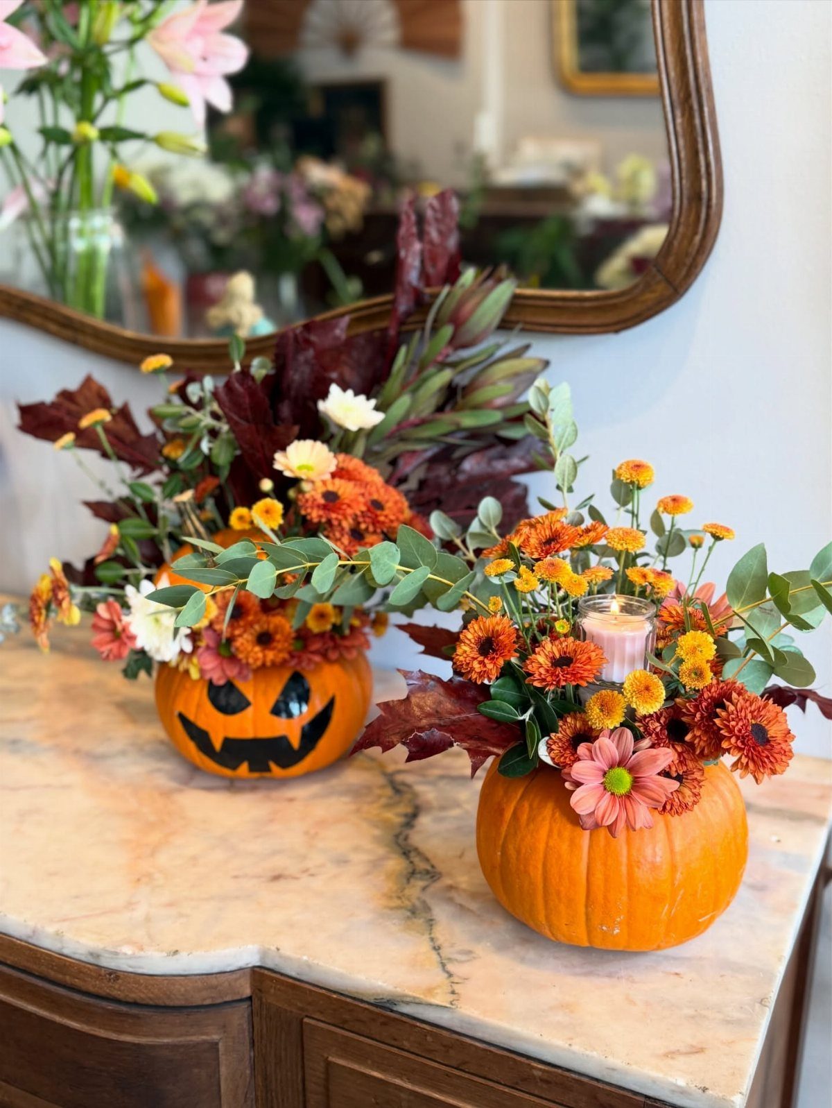 Two orange pumpkin vases filled with autumn flowers on a marble countertop.