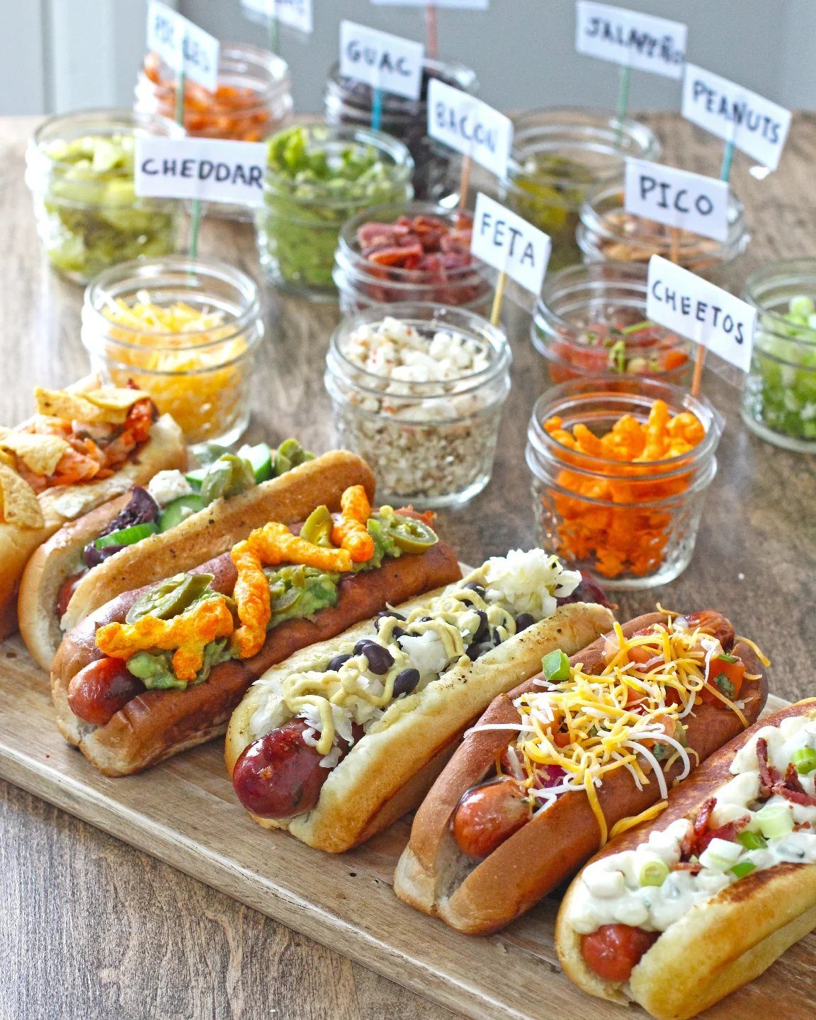 Gourmet hot dogs with diverse toppings and condiment jars on a wooden board.