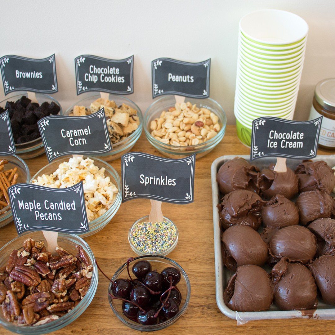 DIY sundae bar with chocolate ice cream, various toppings in bowls, and labels.