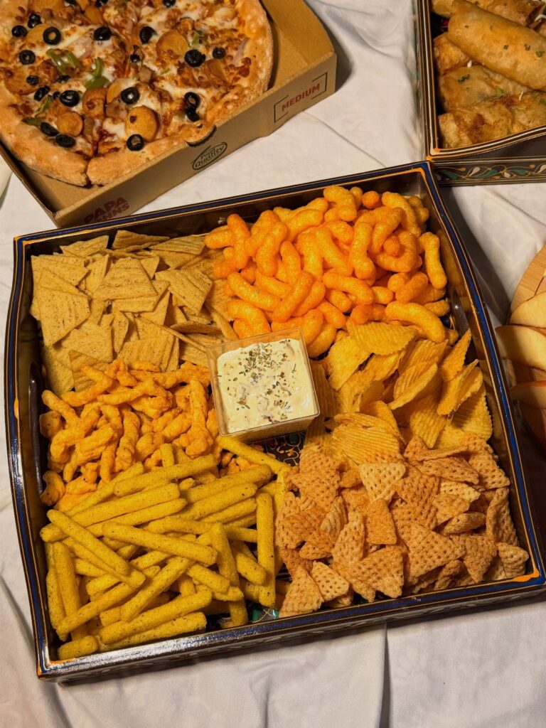Assorted savory snacks in a black tray with pizza and spring rolls nearby.