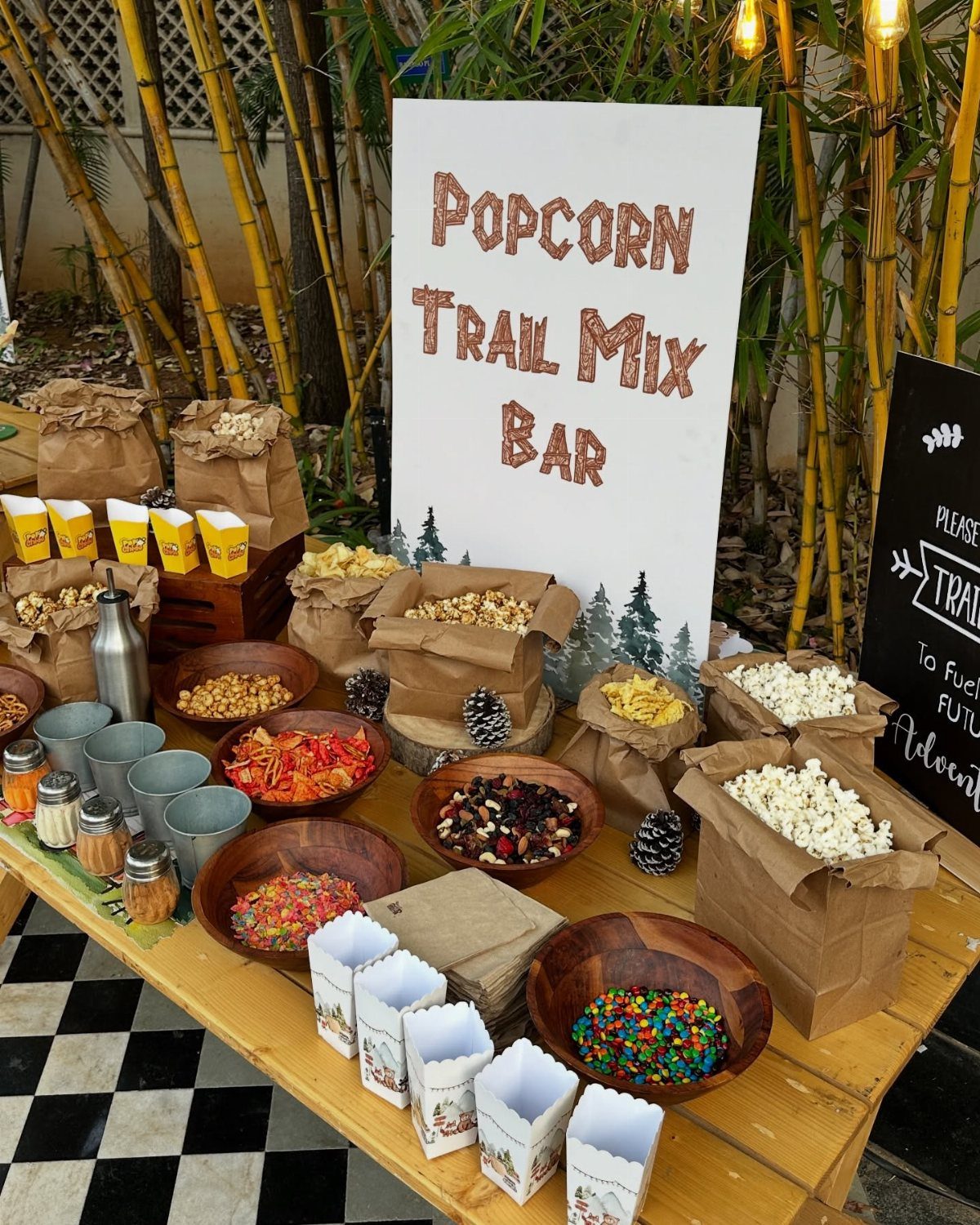 Popcorn trail mix bar with various toppings in bowls and paper bags on wood.
