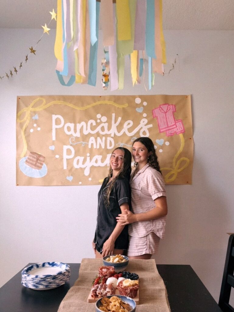 Two women in pajamas posing before a Pancakes and Pajamas party banner and food.