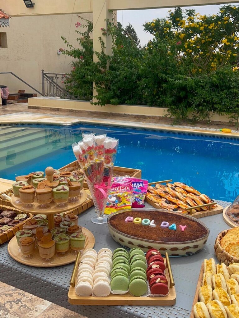 Assorted party snacks and a Pool Day cake on a table by a pool.