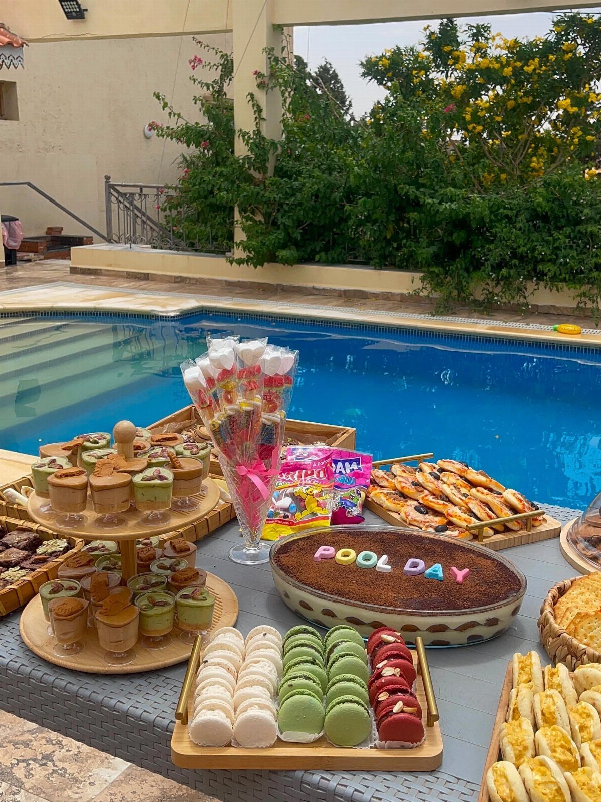 Assorted party snacks and a Pool Day cake on a table by a pool.