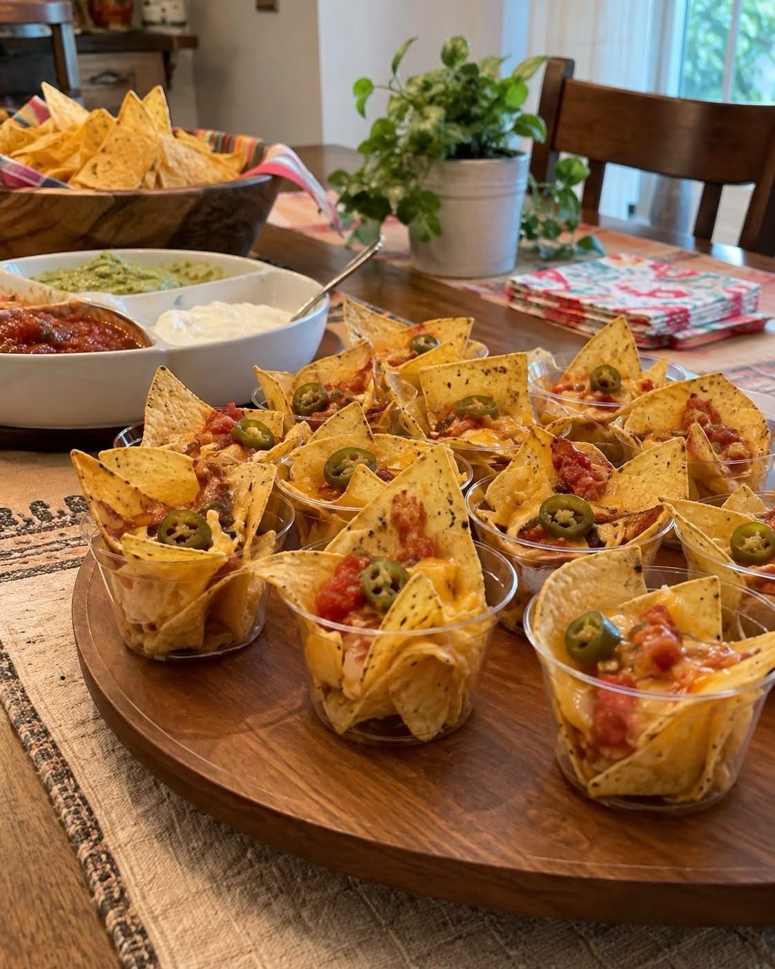 Nachos in small plastic cups on a wooden platter with various dips nearby.