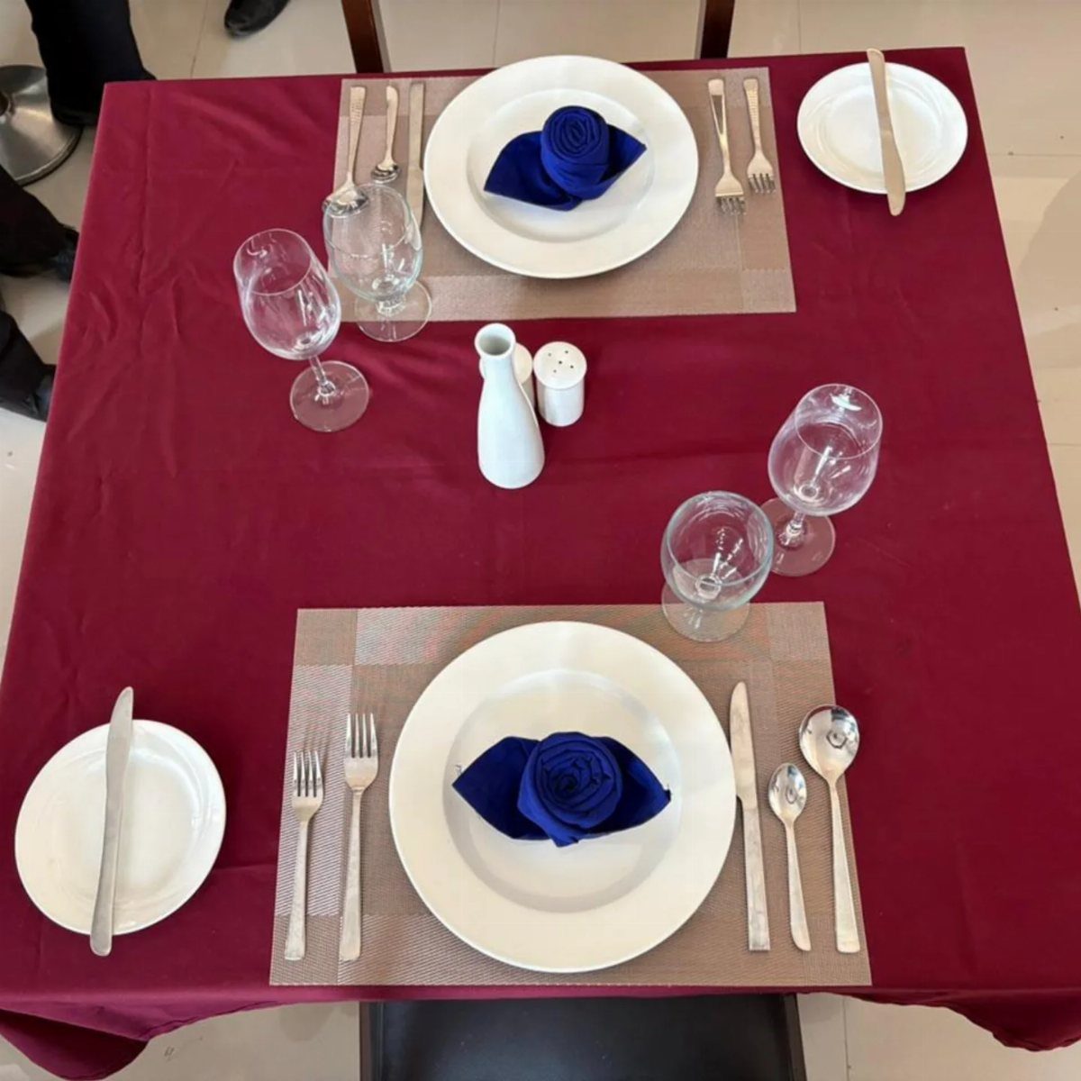 Dinner table set for two with burgundy cloth, white plates, and blue napkins.