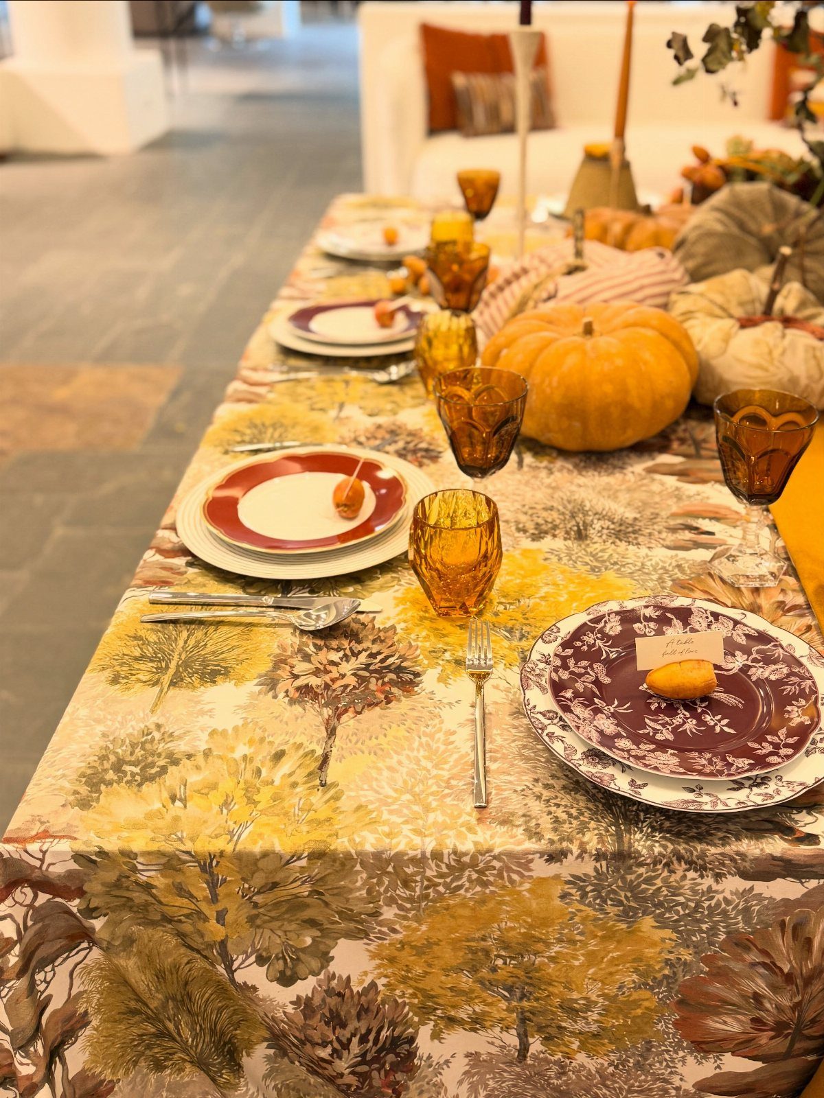 Autumnal table set up with forest-patterned cloth, amber glasses, and orange pumpkins.