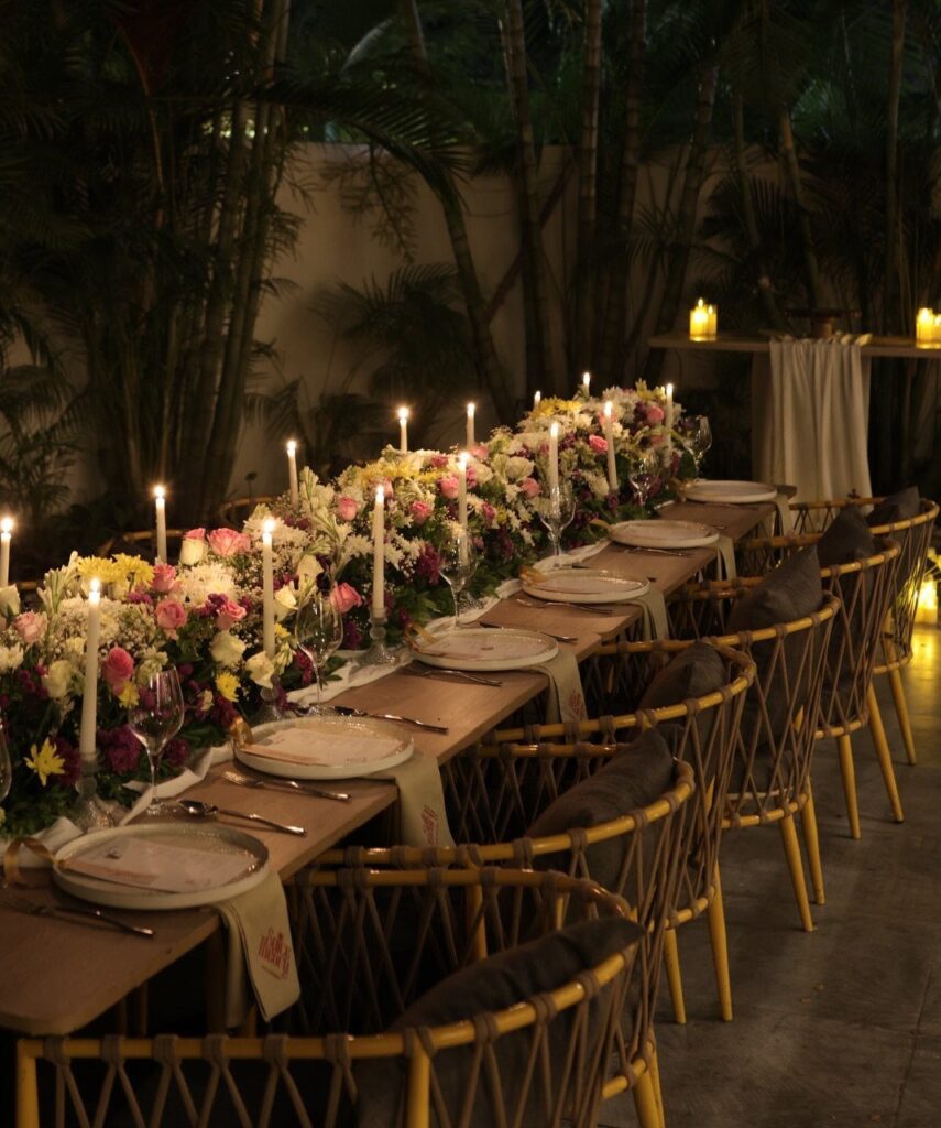 Long dinner table with floral runner, lit candles, and elegant place settings outdoors.