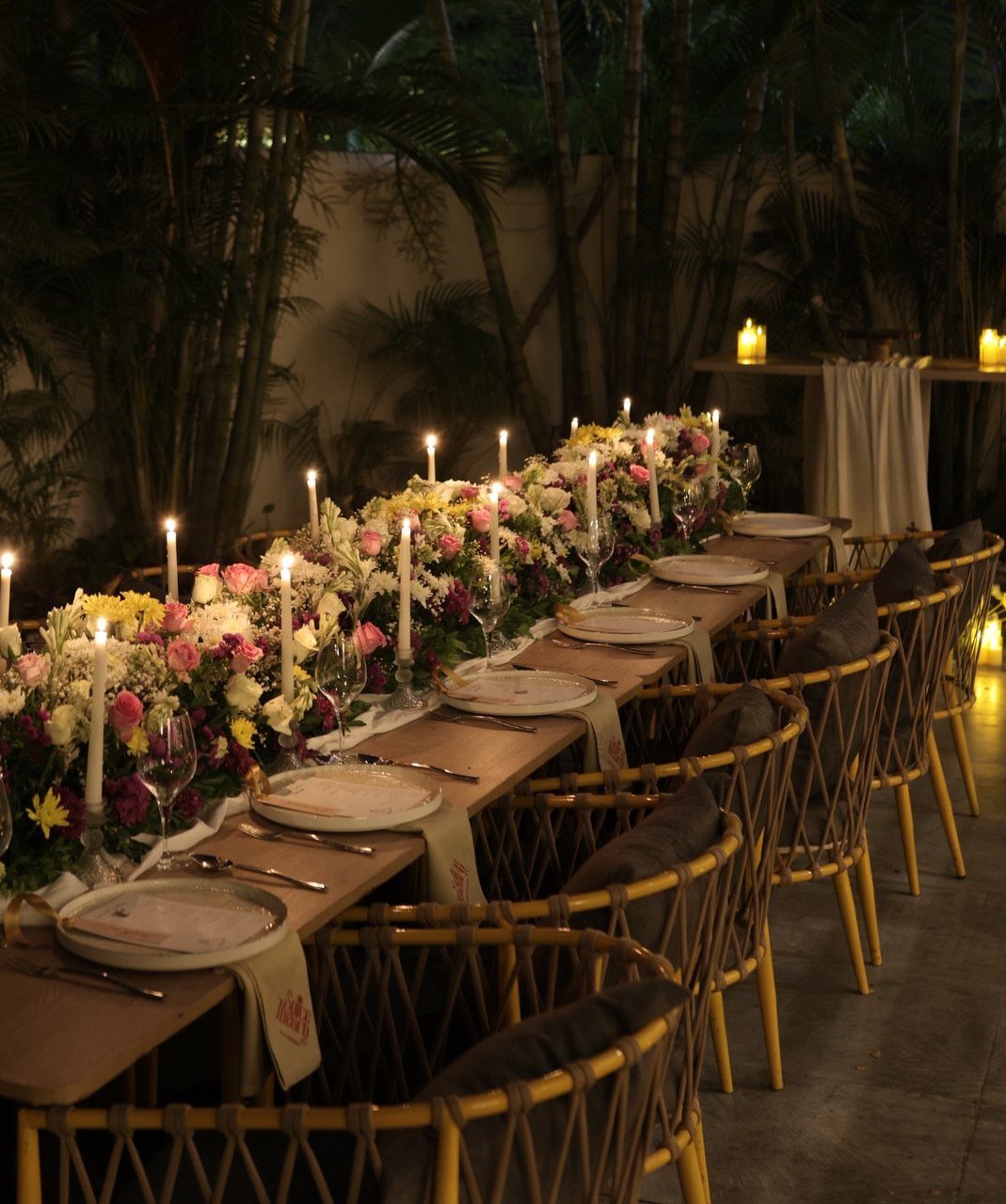 Long dinner table with floral runner, lit candles, and elegant place settings outdoors.