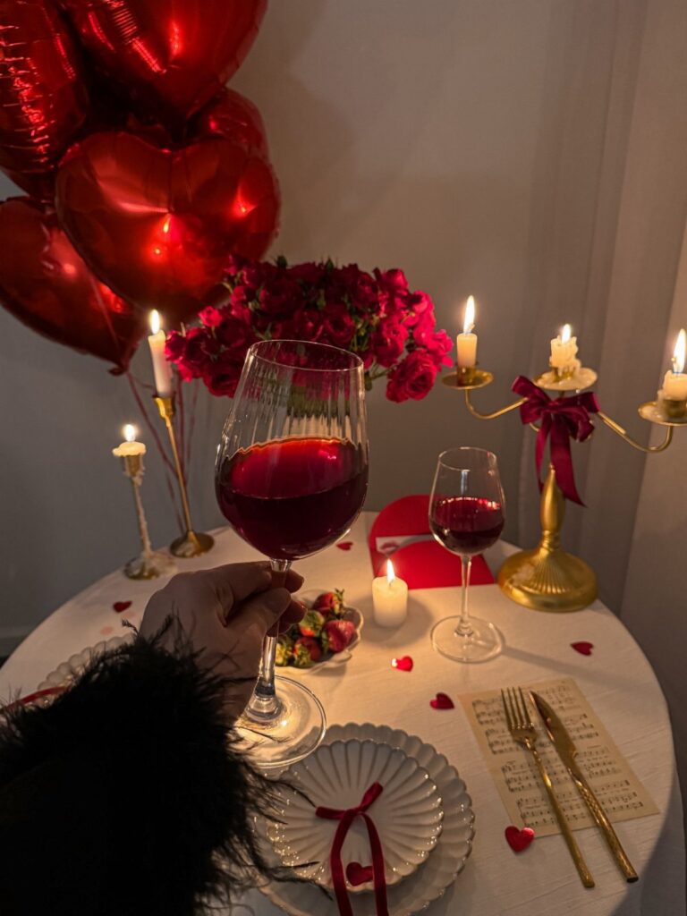 Romantic dinner table with red wine, roses, candles, and heart-shaped balloons.
