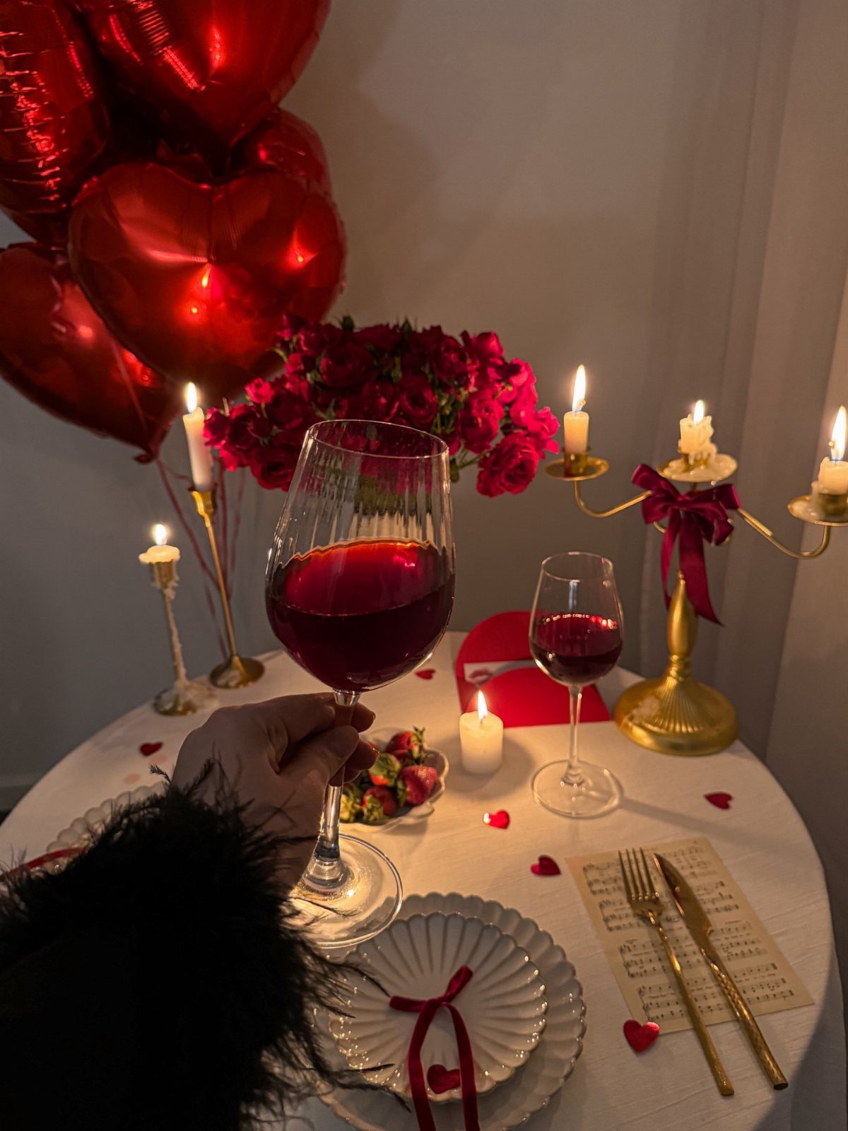 Romantic dinner table with red wine, roses, candles, and heart-shaped balloons.